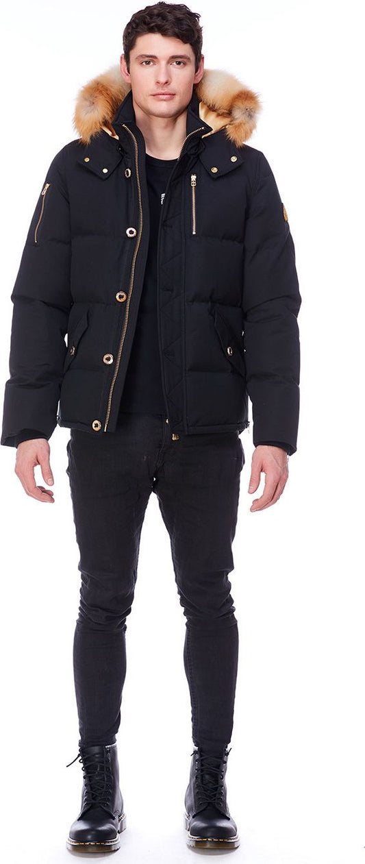 A man stands wearing a black puffer jacket with a fur-lined hood, black pants, and black boots.