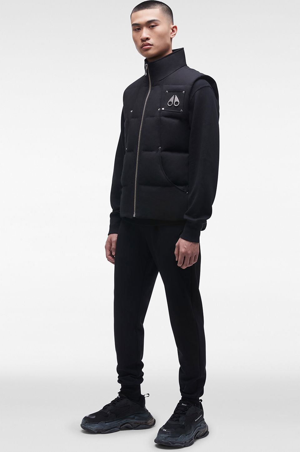 A slim Asian man wearing a black down vest over a black long-sleeve shirt with black pants and black shoes. The vest has a zipper down the front and a small logo on the upper left chest.