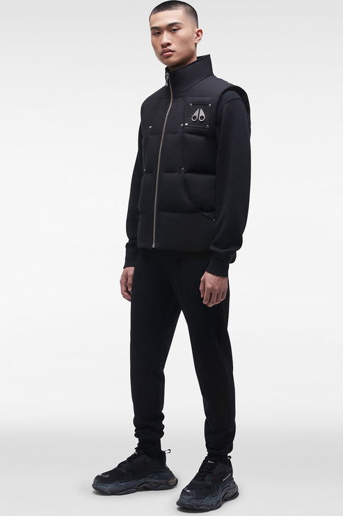 A slim Asian man wearing a black down vest over a black long-sleeve shirt with black pants and black shoes. The vest has a zipper down the front and a small logo on the upper left chest.