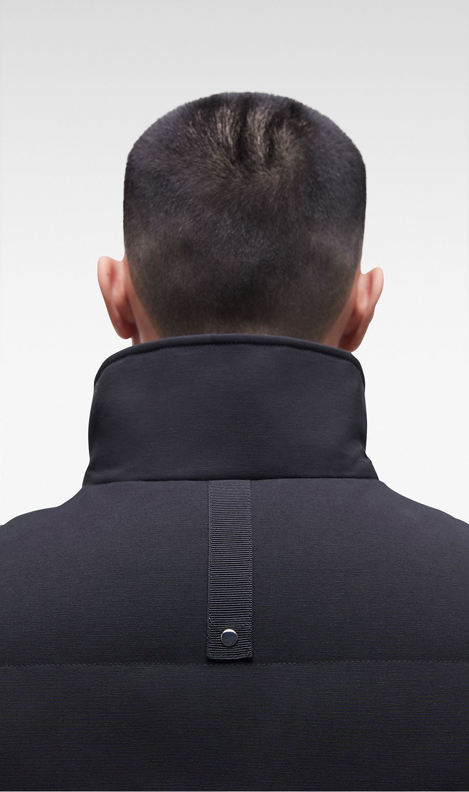 Man wearing a black jacket, back view. The jacket has a stand-up collar and a vertical strap on the back with a button.