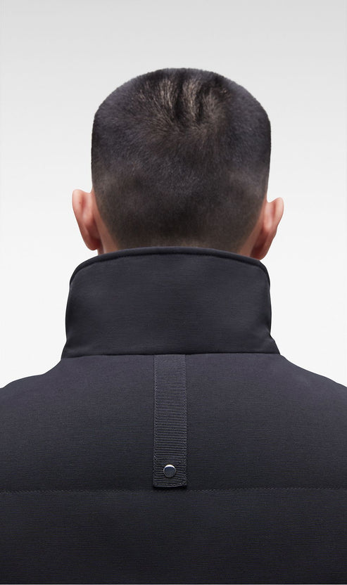 Man wearing a black jacket, back view. The jacket has a stand-up collar and a vertical strap on the back with a button.