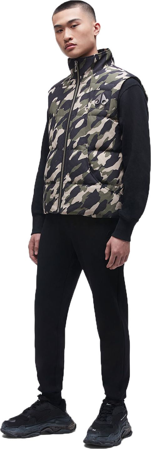 A person wearing a camouflage vest, black long sleeve shirt, black pants, and black sneakers.
