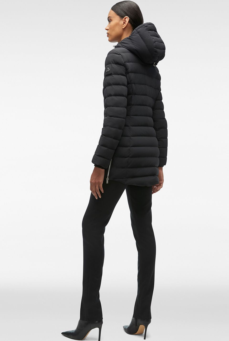 A woman is standing, wearing a black puffy coat with a hood and black pants with black heels.