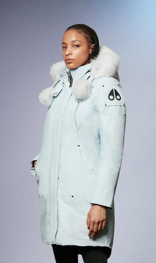 A fair-skinned woman in a light blue winter coat with a white fur-lined hood and the moose knuckles logo on the left shoulder.