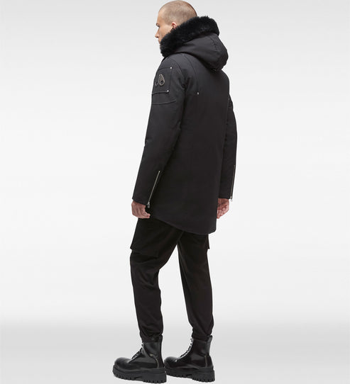 A man in profile wearing a black parka with a fur-trimmed hood, black pants, and black boots.