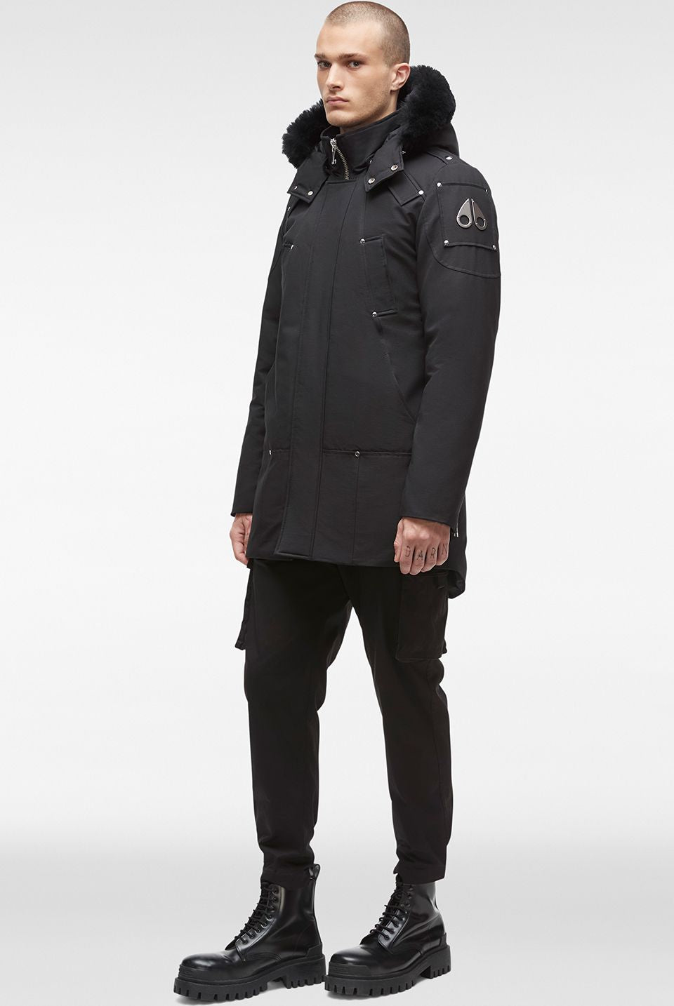 Man in a black parka with fur trim, black pants, and black boots, standing.