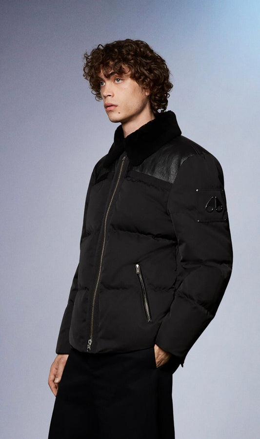 A man with curly hair wearing a black puffer jacket with a black fur collar and leather shoulder patches.