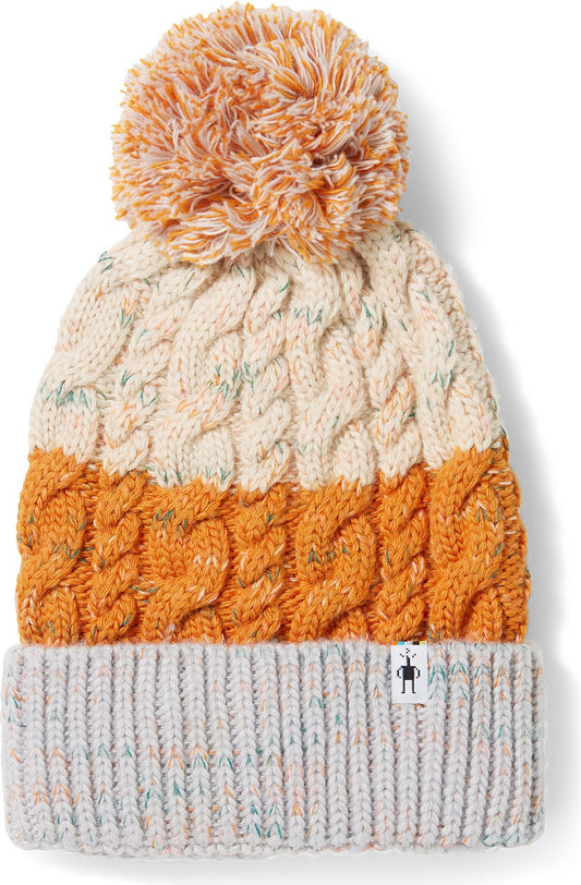Close-up of a winter beanie with a pompom on top, featuring a cable knit design and color blocking in white, orange, and light gray.