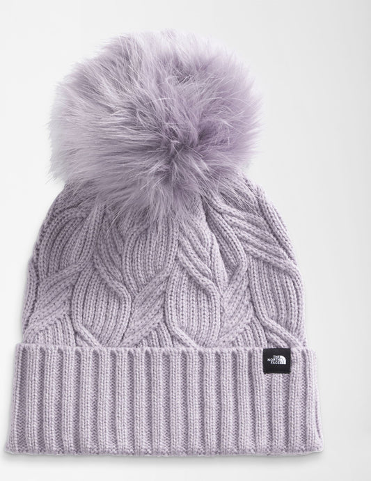 Knitted light purple beanie with a large pompom on top, ribbed brim, and a small black square tag.