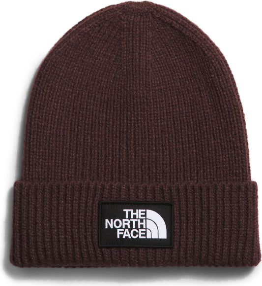 Close-up of a knit beanie in solid dark brown with a thick folded brim and a black square logo patch with white text and a white half-dome symbol.
