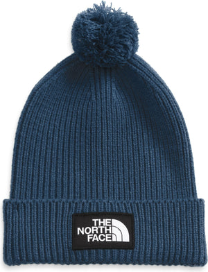 Close-up of a blue knit beanie with a pompom and a black patch with the words "THE NORTH FACE" written in white.