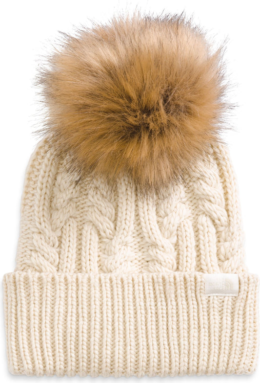 Cream-colored knitted cable beanie with a large faux fur pompom and a small white label near the ribbed brim.