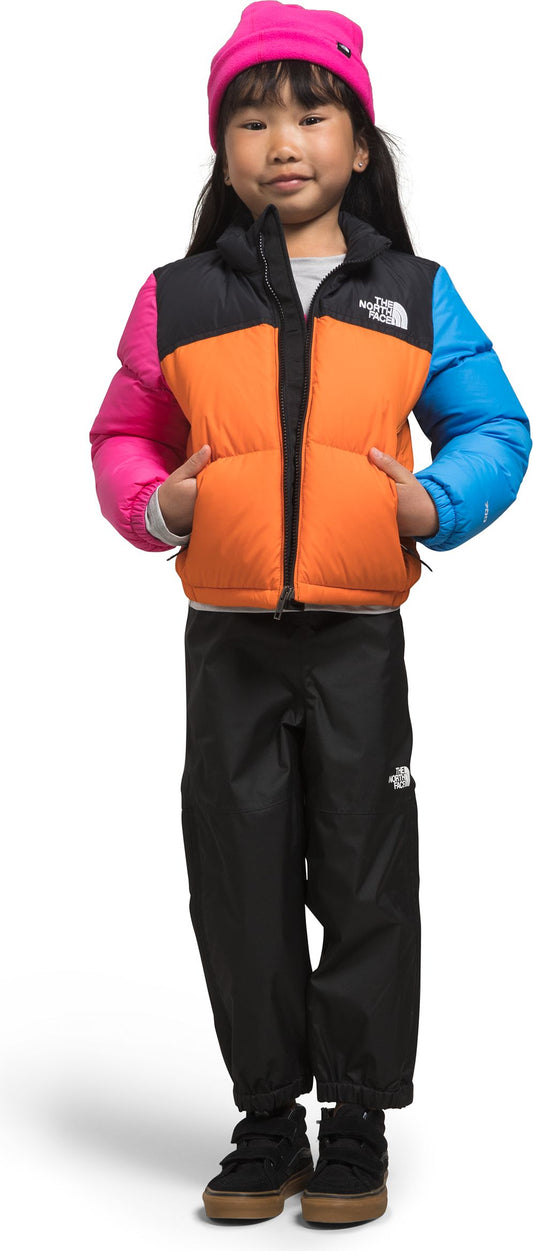 A child wearing a pink beanie, color-blocked puffer vest, black snow pants, and brown sneakers.
