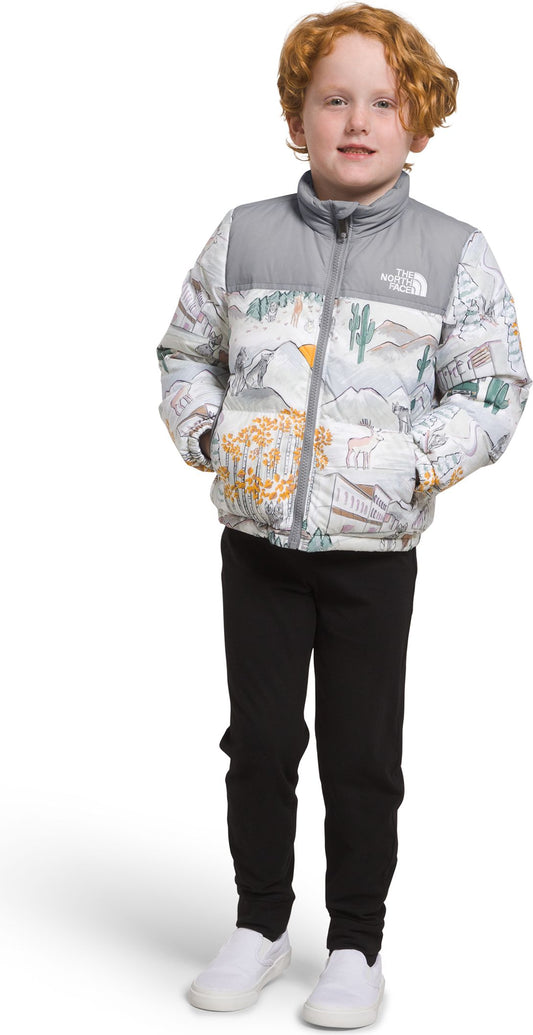 A young boy with red hair and pale skin is wearing a patterned white, gray, and yellow puffer jacket with his hands in his pockets. He is also wearing black pants and white shoes.