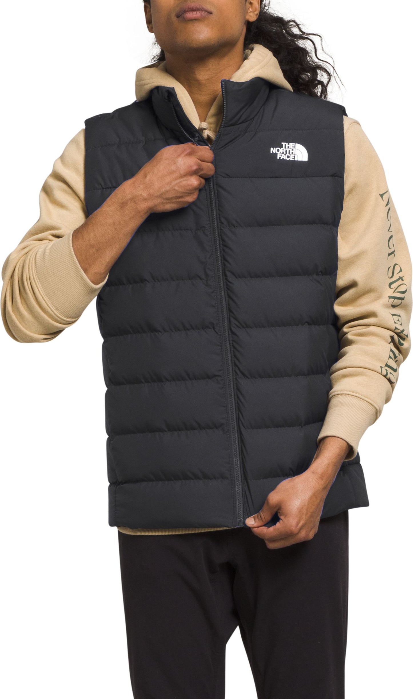 A person wearing a black puffy vest over a tan hoodie, and black pants.