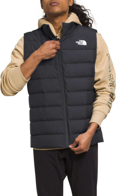A person wearing a black puffy vest over a tan hoodie, and black pants.