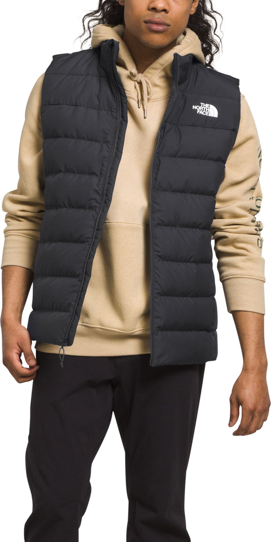 A man wearing a black puffer vest over a tan hooded sweatshirt and black pants.