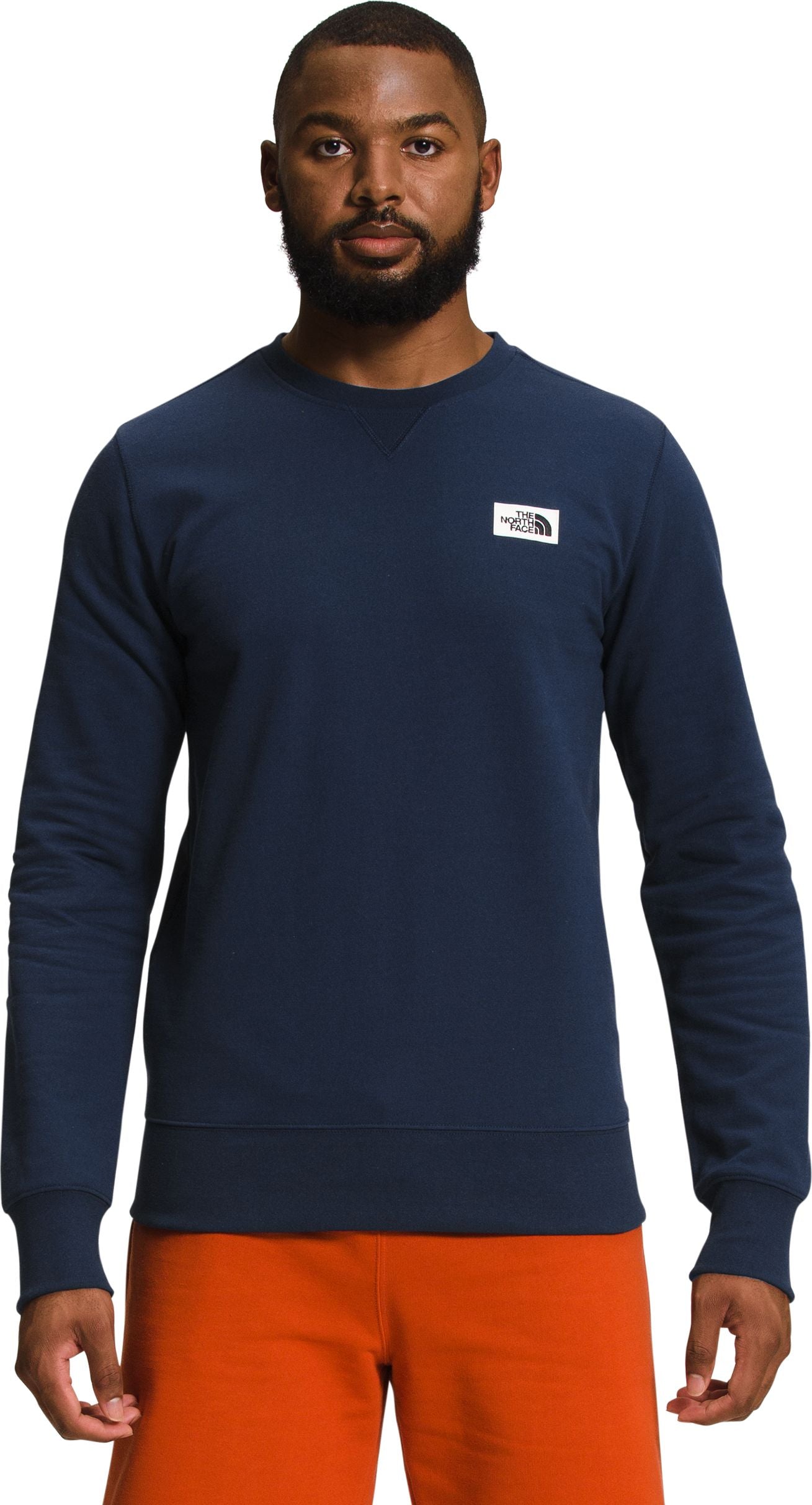 Man in navy crewneck sweatshirt with a logo patch on the upper left chest, and orange shorts.