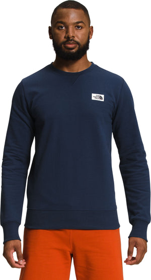 Man in navy crewneck sweatshirt with a logo patch on the upper left chest, and orange shorts.