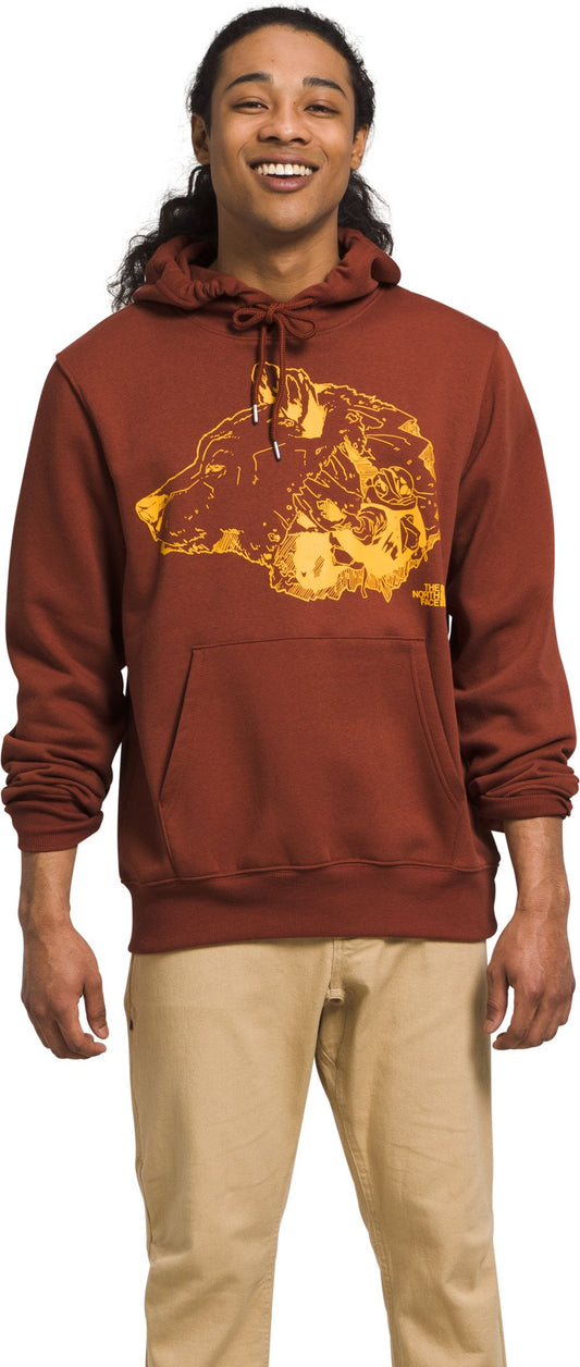 A man wearing a brown hoodie with a yellow bear design and beige pants smiles.