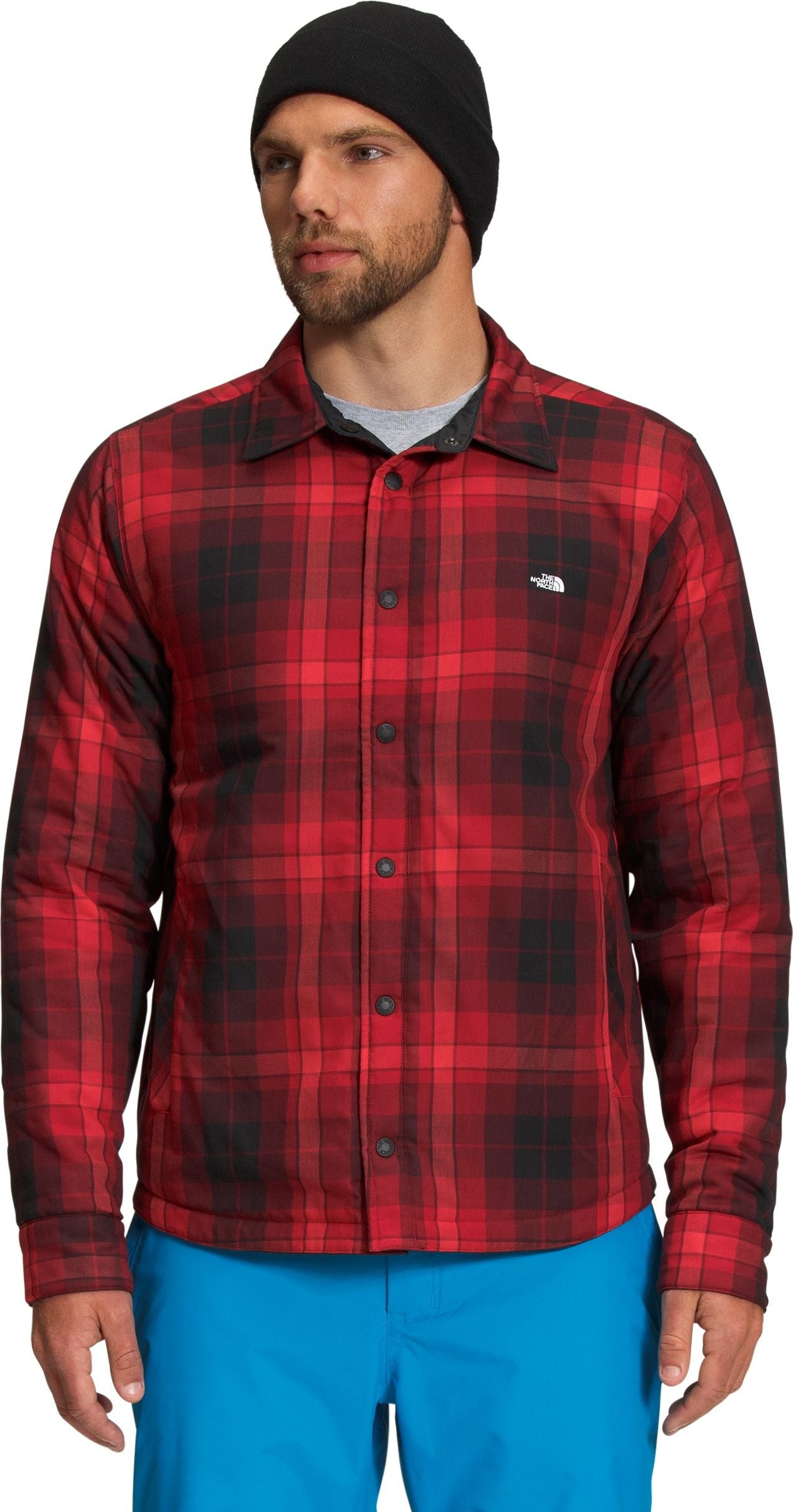 A fair-skinned man wearing a black beanie and a red and black plaid collared shirt with long sleeves and a white logo on the upper right.