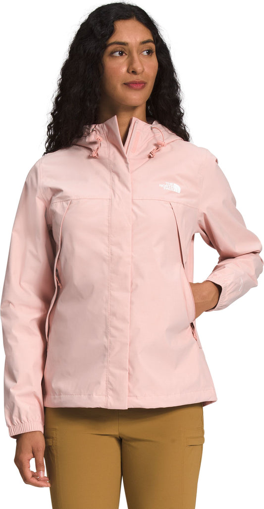 A woman with dark curly hair wears a light pink hooded jacket and tan pants.