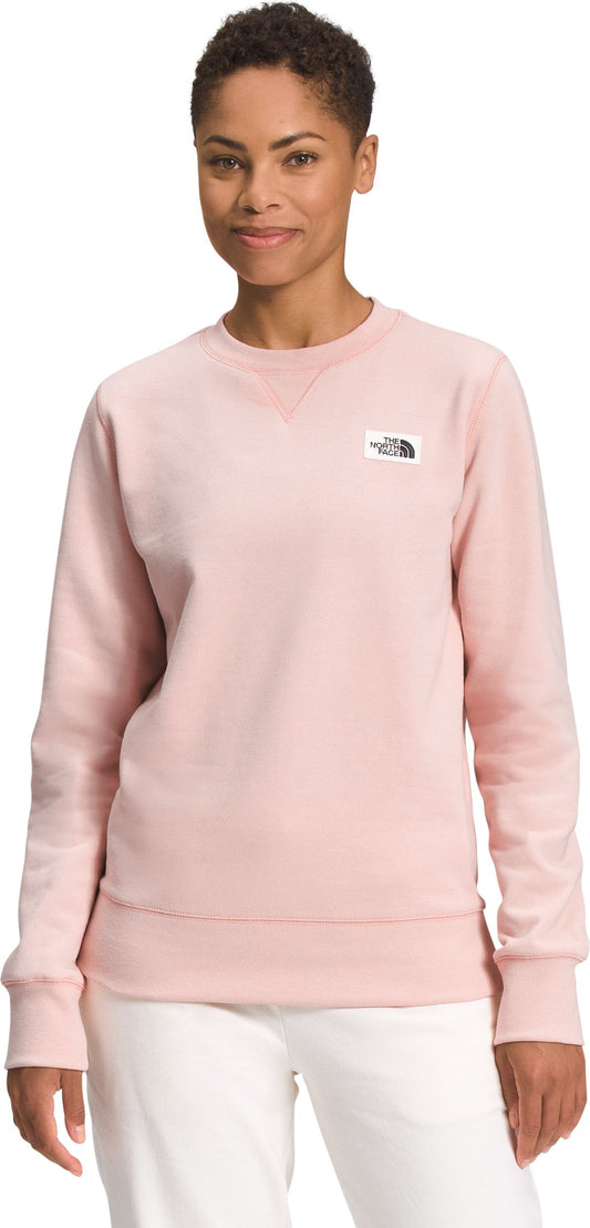 A woman wearing a light pink sweatshirt and white pants is looking at the camera.