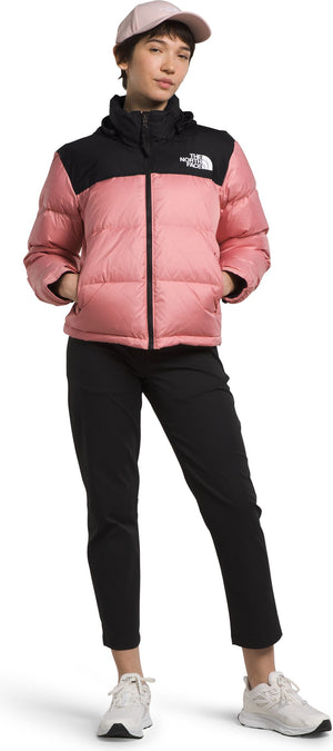 A woman in a pink baseball cap, pink and black jacket, black pants, and white shoes stands in front of a white background.
