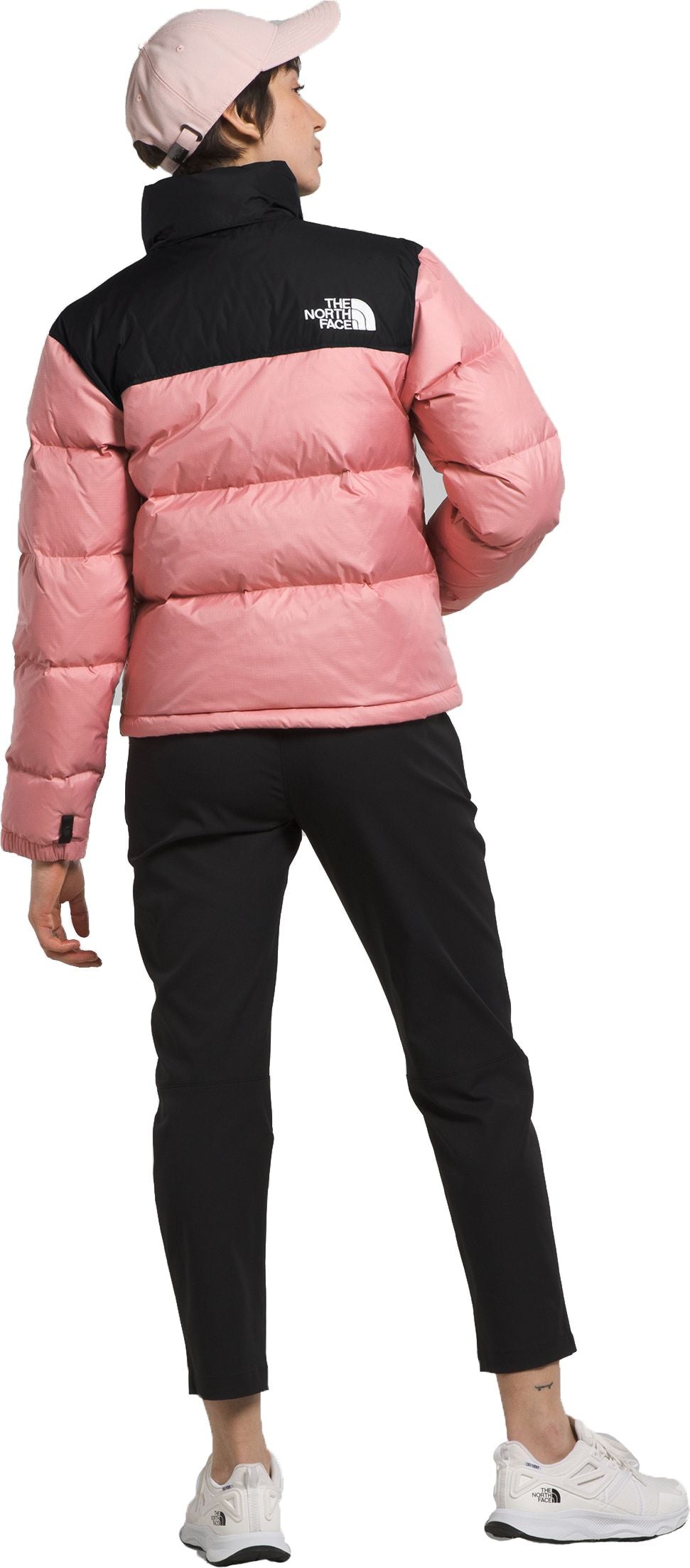 A person wearing a pink cap and a pink and black puffer jacket with black pants and white sneakers, viewed from the back.