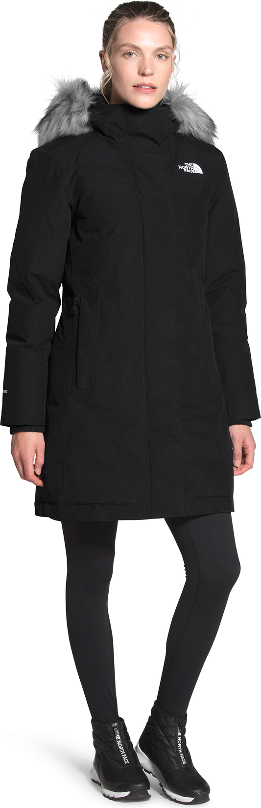 A woman in a long black coat with fur trim, black leggings, and black sneakers.
