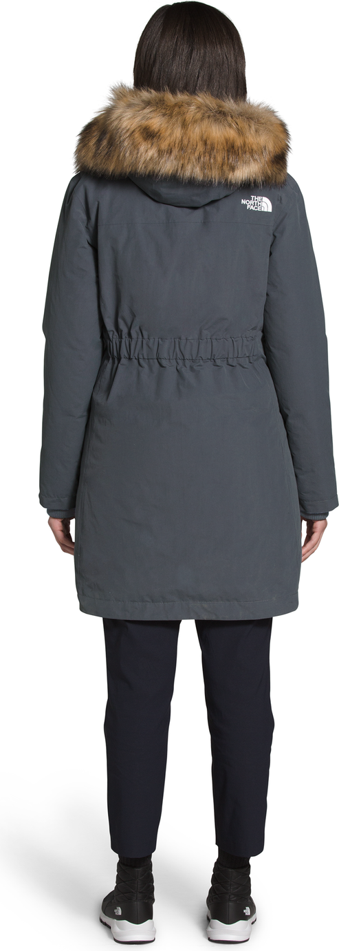 A woman wearing a gray parka with a fur-trimmed hood and dark pants and shoes, facing away from the viewer.