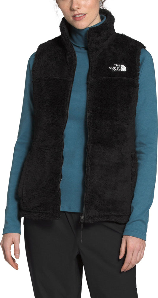 A person wearing a black, fuzzy, sleeveless vest over a teal turtleneck and black pants.