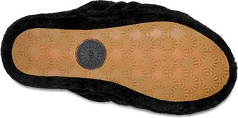 A close-up shows the bottom of a black slipper with a round, dark gray UGG logo and a tan sole covered in a pattern of small sun-like shapes.